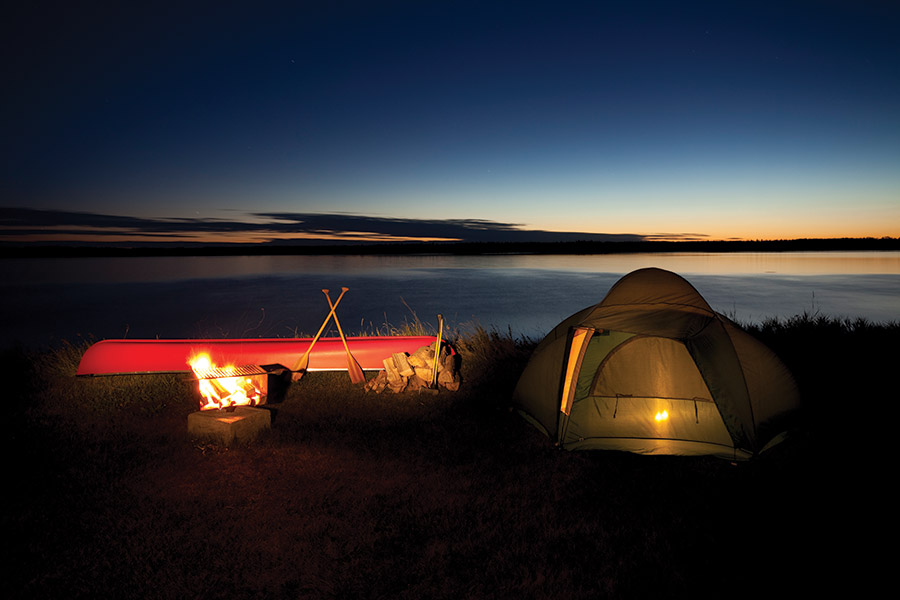 Camping on the shore of Lake Audy in Riding Mountain National Park, Manitoba, Canada. A red canoe and a lit campfire are pictured beside a yellow pop tent at sunset.