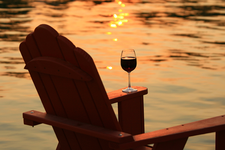A glass of red wine on the arm of an adirondack chair near the calm water of a lake in the Parkland